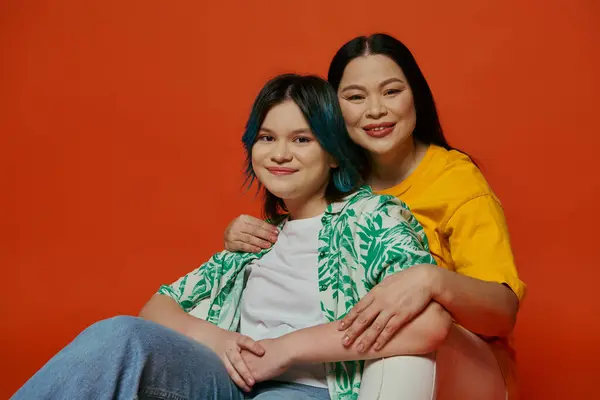 stock image An Asian mother and her teenage daughter sit gracefully on a chair against a vibrant red background.