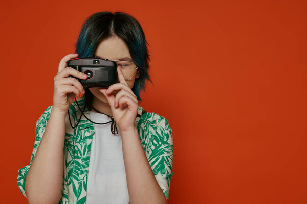Asian teen girl holds a camera up to her face in a studio on an orange background.