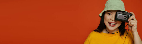 An Asian woman holds a camera to her face in a studio against an orange backdrop.
