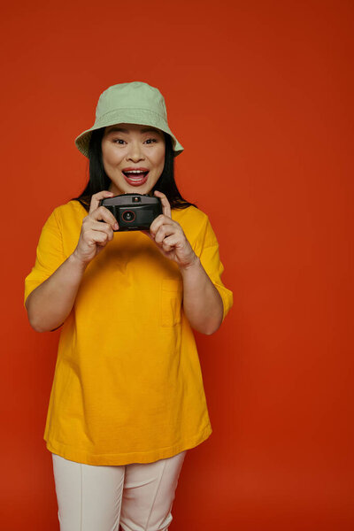 An Asian woman in a yellow shirt captures moments with a camera on in a studio on an orange background.
