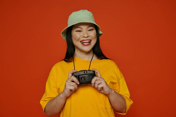 An Asian woman happily holds a camera, smiling at the viewer, in a studio