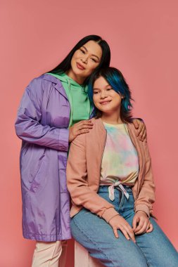 Asyalı bir anne ve genç kızı, canlı pembe bir arka plana karşı günlük giysiler içinde yan yana otururlar..