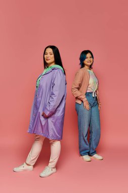 Asyalı bir anne ve genç kızı, pembe bir arka planda günlük giysiler içinde yan yana dururlar..