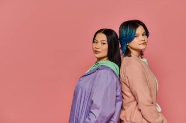 A stylish Asian mother and her teenage daughter, both with vibrant blue hair, pose together on a pink studio background. clipart