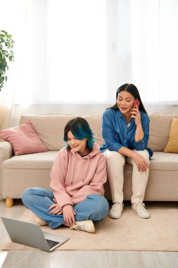 Asyalı anne ve kızı gündelik giysiler içinde, bir kanepede oturmuş, ayrı dijital dünyayla meşgul.