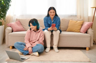 Asian mother and her teenage daughter, wearing casual attire, are sitting on a couch and focused on a cell phone screen. clipart