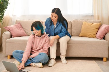 Asian mother and her teenage daughter sitting on a couch, focused on a laptop screen in a cozy home setting. clipart