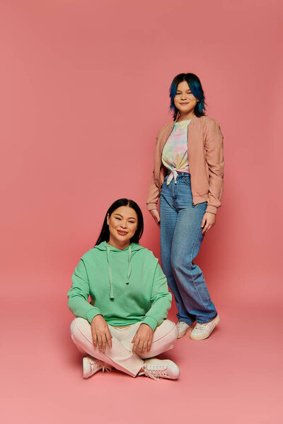 Asian mother and her teenage daughter, dressed casually, standing together against a pink background in a studio setting.