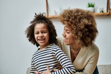 Mutlu bir Afrikalı Amerikalı anne ve kız evlerinde kanepede otururken birlikte gülüyorlar..