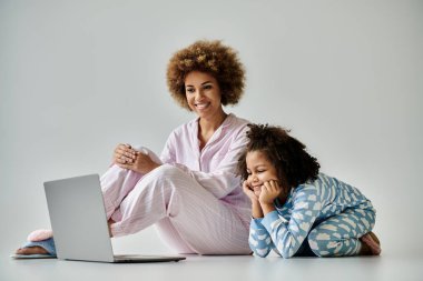 Pijamalı mutlu bir Afrikalı anne ve kız yerde dizüstü bilgisayar kullanarak oturuyor..