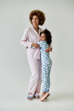 Mutlu Afrika kökenli Amerikalı anne ve kız pijama giyip gri bir arka planda kucaklaşıyorlar..