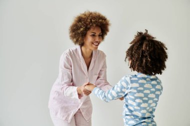 Pijamalı Afrikalı bir anne ve kız el sıkışıyor. Gri bir arkaplanda sıcaklık ve birlik yayıyorlar..
