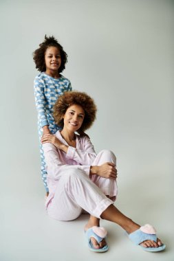 A cheerful African American mother and daughter in matching pajamas strike a pose on a grey background. clipart