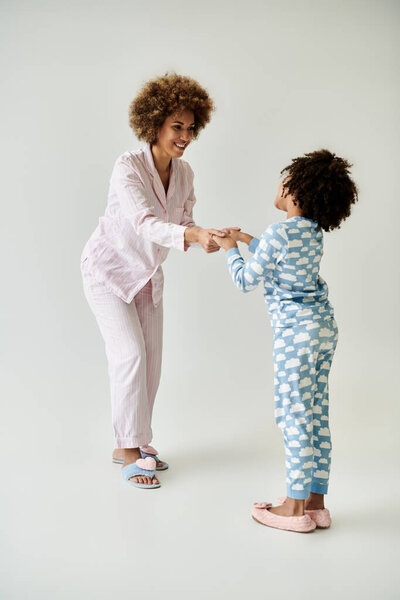 A happy African American mother and daughter in matching pajamas share a warm moment together on a grey background.