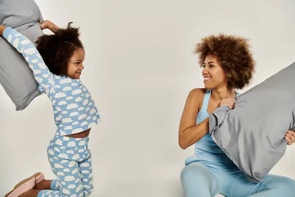 stock image African American mother and daughter in pajamas playfully tossing pillows in the air on a white background.