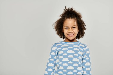 Genç bir Afro-Amerikalı kız mutlu bir şekilde bulutlu mavi bir gömlek giyiyor, gri bir arka planda poz veriyor..