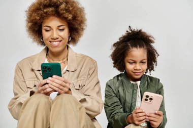 Şık giyinmiş kıvırcık saçlı Afrikalı bir anne ve kız dijital dünyalara dalmış telefonlarına odaklanıyorlar..