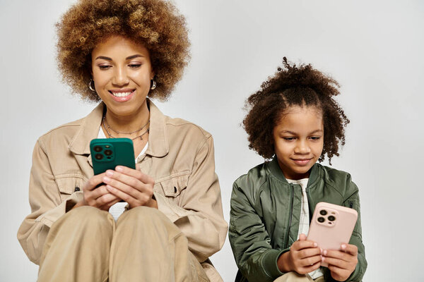 A curly African American mother and daughter in stylish clothes focus on their phones, engrossed in digital worlds.