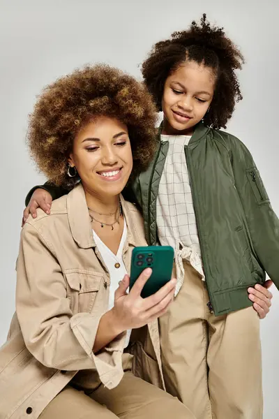 stock image A curly African American mother and daughter, stylishly dressed, engrossed in a cell phone on a grey background.