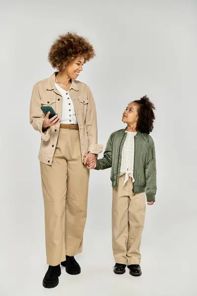 stock image Curly African American mother and daughter in stylish attire, holding hands against a grey backdrop.