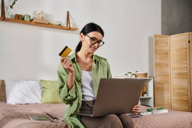 Yatakta oturan bir kadın, elinde bir kredi kartı ve dizüstü bilgisayarla uzaktan çalışıyor..