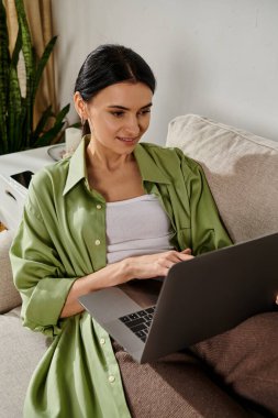 Gündelik giyinmiş bir kadın bir koltukta oturur, dizüstü bilgisayar kullanmaya kendini kaptırmış bir şekilde..