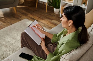 Kanepede oturan, gündelik kıyafetli bir kadın, bir deftere gömülmüş..