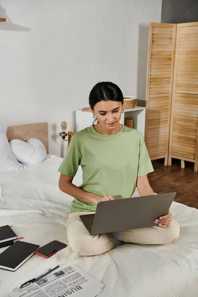 stock image A woman in casual attire sitting on a bed, typing on a laptop computer.