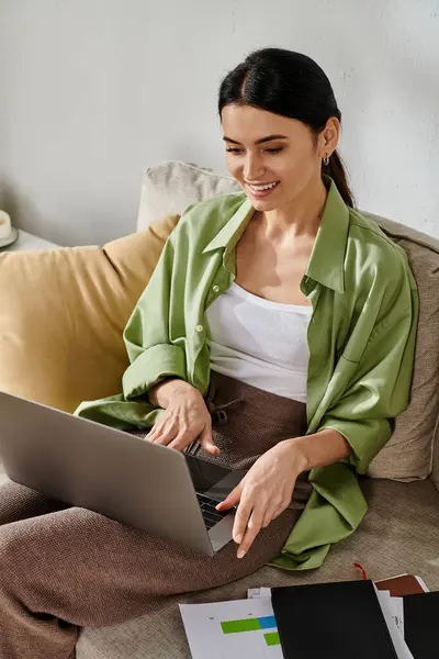 stock image A woman in casual attire works remotely on a laptop computer while seated on a couch.