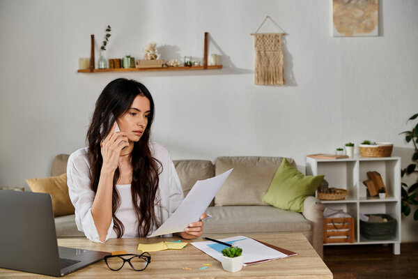 A woman in casual attire works on her laptop at a table with papers.