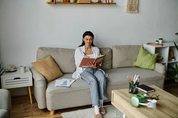 stock image Woman in casual attire sitting on a couch immersed in reading a book.