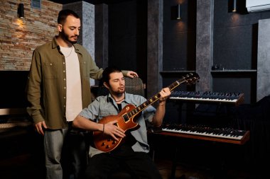 İki adam yan yana durur, bir müzik grubu provasına dalmış bir şekilde kayıt stüdyosunda.
