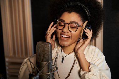 Bir müzik grubu provası sırasında, gözlüklü ve afro saçlı bir kadın mikrofona tutkuyla şarkı söylüyor..