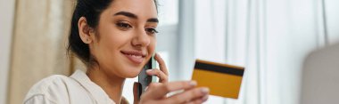 Sıradan giyinmiş bir kadın elinde kredi kartıyla telefonuyla konuşuyor..