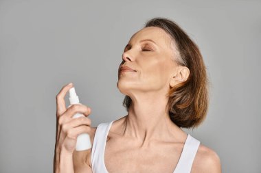 A mature woman in comfy attire is actively using a spray on her face for a refreshing moment. clipart