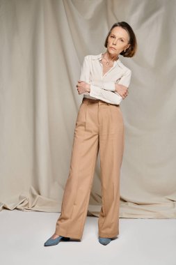 Beyaz bluz ve kahverengi pantolon giyen olgun bir kadın zarif bir poz verir..