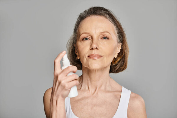 Стильная женщина в повседневной одежде с бутылкой спрея на сером фоне.