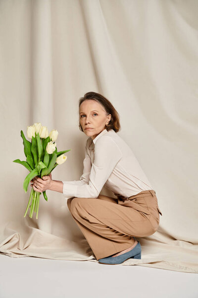 Elegant woman in casual attire crouching, holding a vibrant bouquet of tulips gracefully.