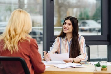 Two women engage in conversation while seated at a desk. clipart