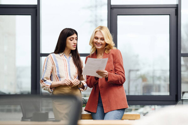 Two women dressed professionally, standing in an office in front of a large window.