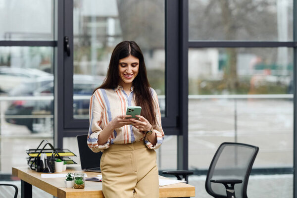 A woman stands at a desk, absorbed in her phone.