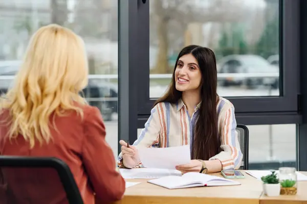 Stock image Two women engage in conversation while seated at a desk.