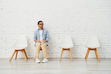A man sits in a row of chairs against a white brick wall. clipart