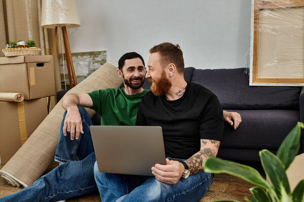 Два влюблённых гея сидят на диване, окружённые движущимися коробками, сосредоточенные на экране ноутбука..