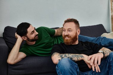 Eşcinsel bir çift, yeni evlerinde taşınan kutularla çevrili bir koltuğa yakın otururken hassas bir anı paylaşıyor..