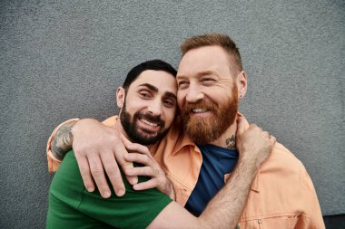 Gri bir duvarın önünde sıcak bir kucaklaşmayı paylaşan, gündelik giysiler içinde eşcinsel bir çift..