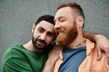Gündelik giyinmiş iki adam, gay bir çift, gri bir duvara karşı sevgiyle dururlar..