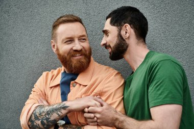 Two men in casual attire stand next to each other, showing affection against a grey wall. clipart