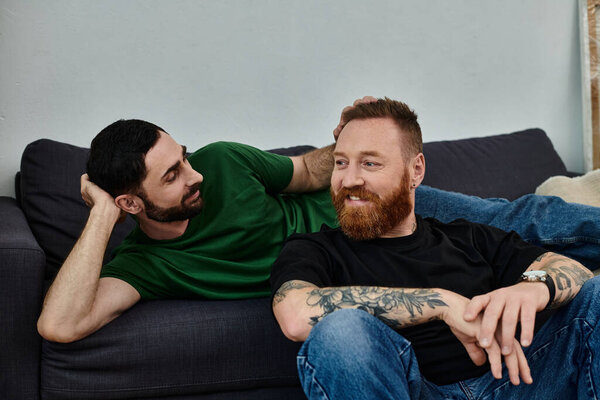 A gay couple shares a tender moment as they sit closely on top of a couch, surrounded by moving boxes in their new home.