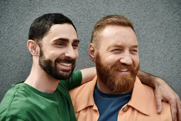 Birbirine aşık eşcinsel bir çift, günlük kıyafetler giyiyorlar, gri bir duvarın karşısında yan yana duruyorlar, aralarındaki güçlü bağı gösteriyorlar..